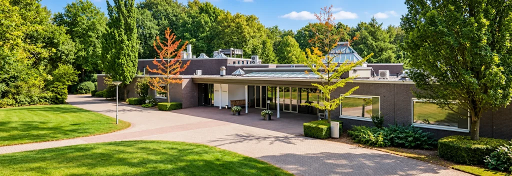 Buitenaanzicht van Crematorium De Meerdijk in Emmen, met de entree van het gebouw, een verzorgde toegangsweg en een groene omgeving met bomen en struiken.