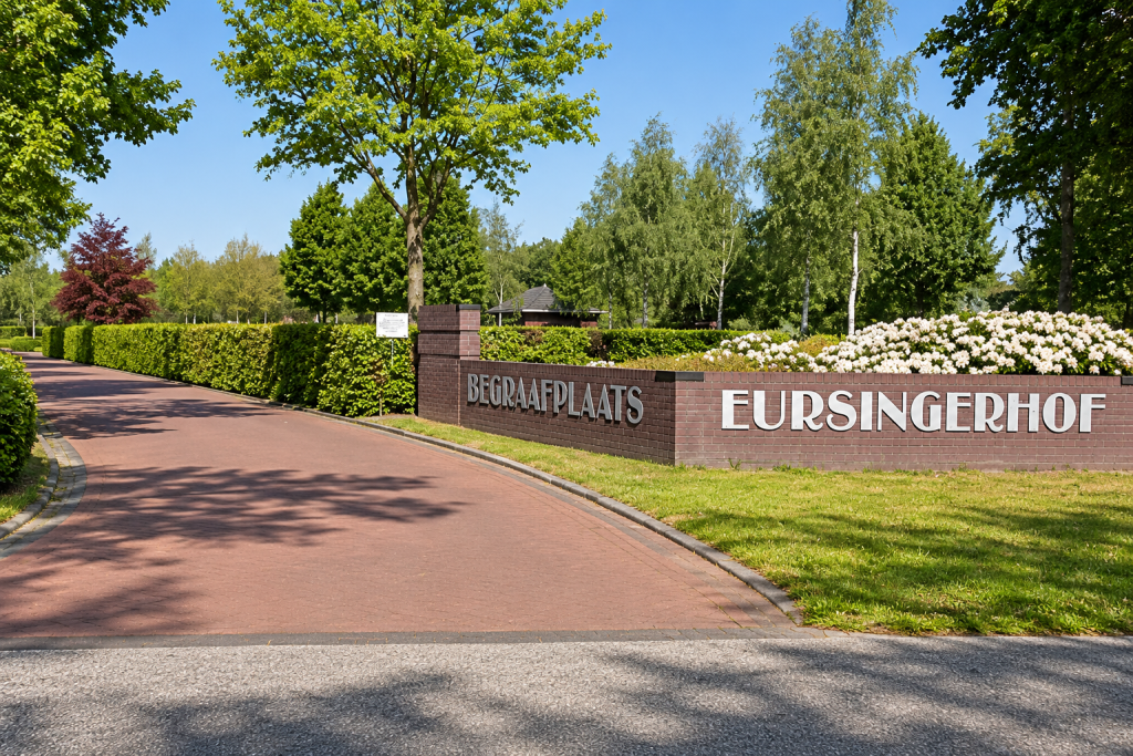 Entree van Begraafplaats Eursingerhof aan de Eursing 4 in Beilen, met een bakstenen naambord, groene hagen, witte bloemen en een verzorgde toegangsweg.