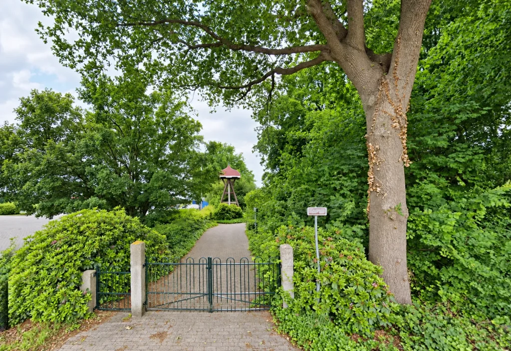 Entree van de Algemene Begraafplaats Nijeveen met een groen toegangshek, een wandelpad, hoge bomen en een houten klokkentoren in een rustige, groene omgeving.