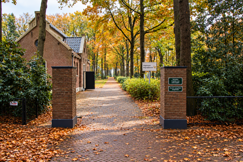 Ingang van de Gemeentelijke Begraafplaats in Hollandscheveld, gemeente Hoogeveen, met een karakteristiek bakstenen gebouw, toegangspoort en een bomenlaan in herfstkleuren.