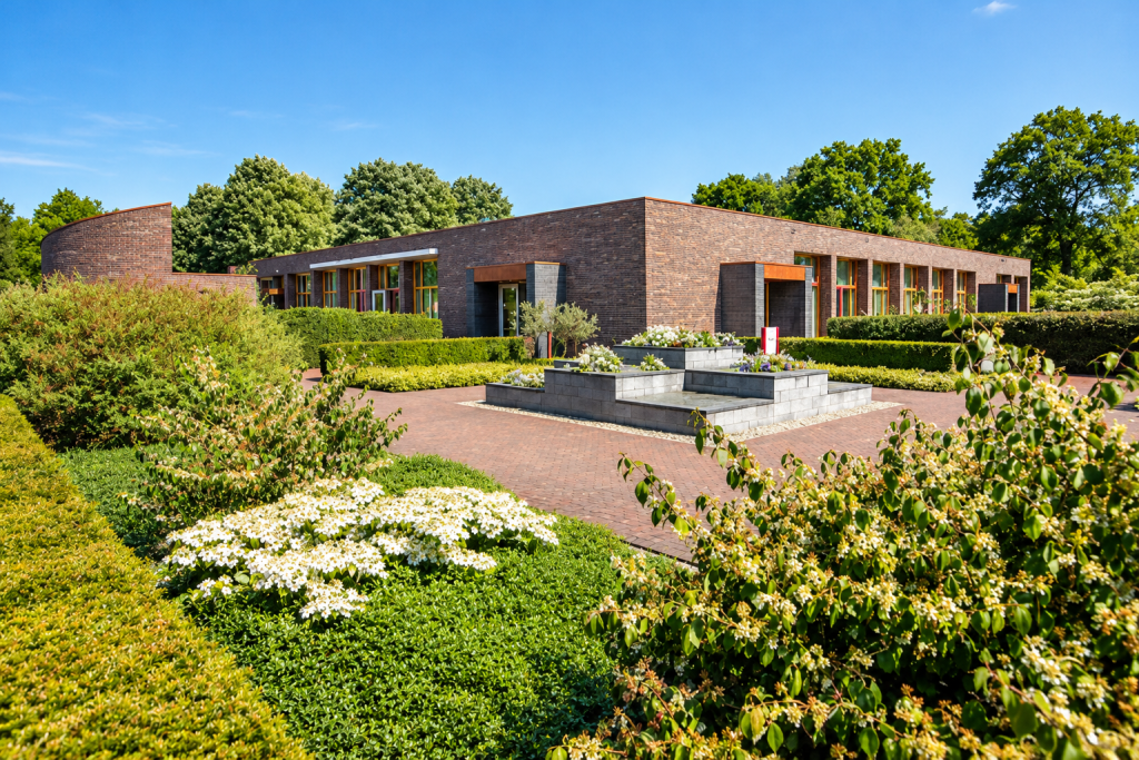 Buitenaanzicht van crematorium Reestborgh bij de Algemene Begraafplaats in Meppel, met een modern bakstenen gebouw, groene tuinen, bloemen en een verzorgde entree.
