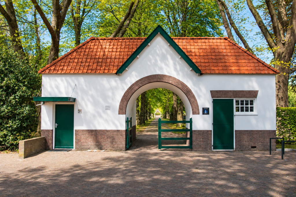 Entree van Begraafplaats Klazienaveen in de gemeente Emmen, met een karakteristiek wit poortgebouw, rood pannendak, groene toegangspoort en een bomenlaan.