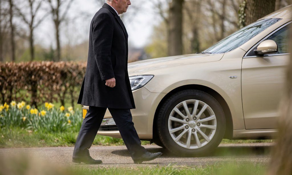 Uitvaart zonder verzekering. Beige rouwauto als onderdeel van een rustige en respectvolle rouwstoet op een landelijke weg, gevolgd door een tweede voertuig, in een natuurlijke en realistische omgeving met zachte daglichtinval en een serene sfeer.