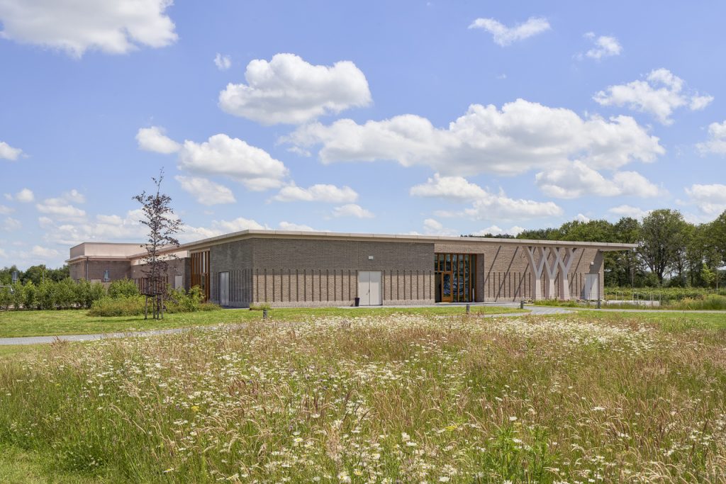Buitenaanzicht van Crematorium Ommeland en Stad, een modern crematorium met een lichte bakstenen gevel, omringd door een groene tuin met wilde bloemen en bomen.
